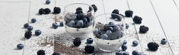Panoramic shot of tasty yogurt with chia seeds, blueberries, blackberries near scattered seeds and berries on white wooden isolated on black — Stock Photo