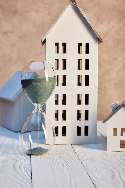 Casas modelos y reloj de arena en mesa de madera blanca cerca de pared texturizada, concepto de bienes raíces - foto de stock