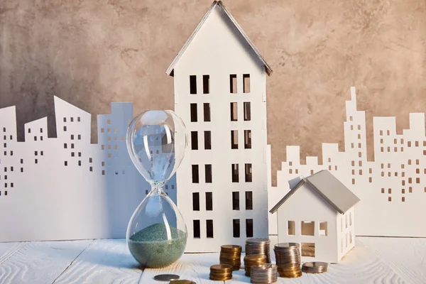 Casas modelos y reloj de arena sobre mesa de madera blanca con monedas, concepto de bienes raíces - foto de stock
