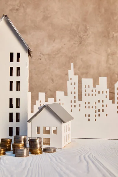 Monedas y casas modelos en mesa de madera blanca cerca de la pared de textura marrón y la ciudad de papel, concepto de bienes raíces - foto de stock