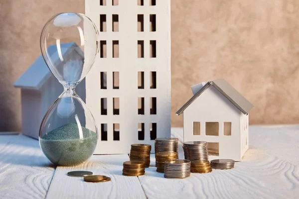 Houses models on white wooden table near hourglass and coins, real estate concept — Stock Photo