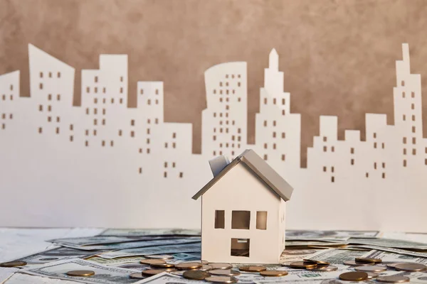 Модель дома на долларовых банкнотах рядом с монетами и бумажным городом на заднем плане, концепция недвижимости — стоковое фото