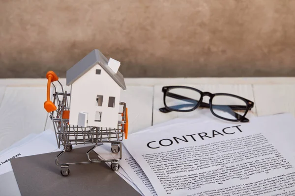 Modelo da casa no carrinho de compras pequeno na mesa de madeira branca com contrato e óculos, conceito imobiliário — Fotografia de Stock