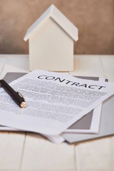 White house model on white wooden table with contract and pen near brown textured wall, real estate concept — Stock Photo