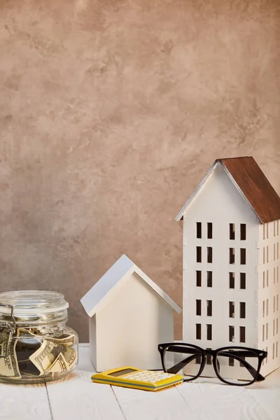 Casas modelos en mesa de madera blanca con caja de dinero, calculadora y gafas cerca de la pared de textura marrón, concepto de bienes raíces - foto de stock