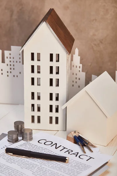 Casas modelos en mesa de madera blanca con contrato, monedas y llaves cerca de pared de textura marrón, concepto de bienes raíces - foto de stock