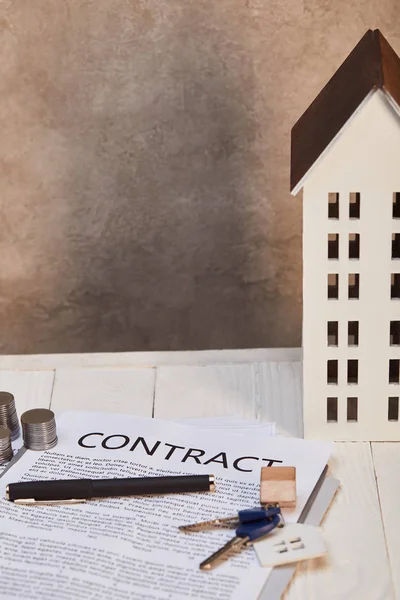 Модель дома на белом деревянном столе с контрактом, монеты и ключи возле коричневой текстурированной стены, концепция недвижимости — стоковое фото