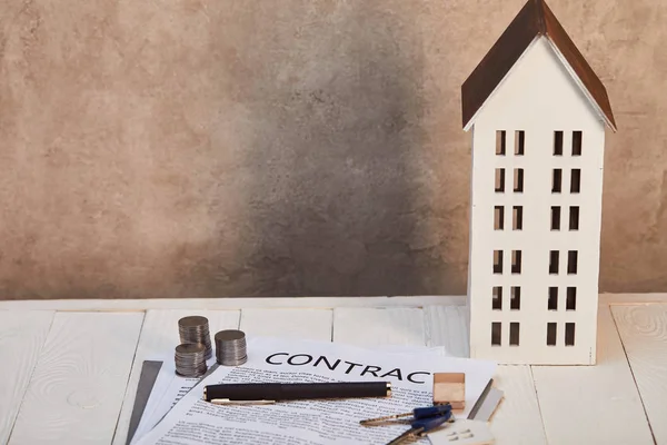 Модель будинку на білому дерев'яному столі з контрактом, монети і ключі біля коричневої стіни, концепція нерухомості — стокове фото