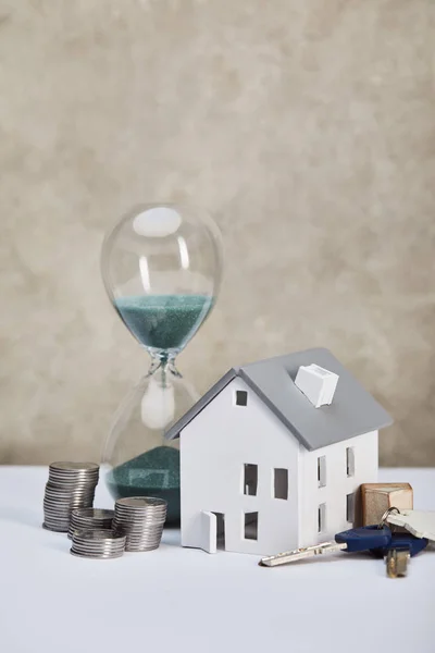 Modelo de casa en mesa blanca con reloj de arena, monedas de plata y llaves, concepto de bienes raíces - foto de stock