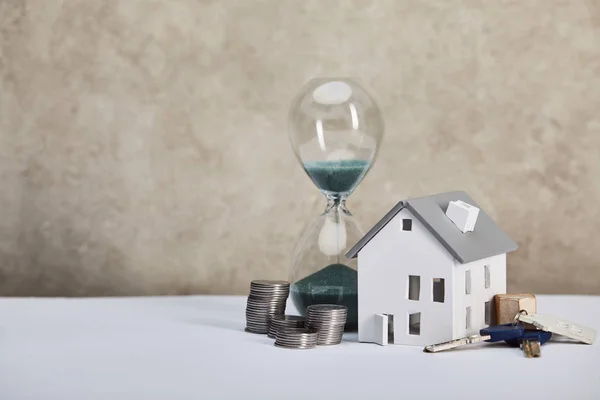 Modelo de casa en mesa blanca con reloj de arena, monedas y llaves, concepto de bienes raíces - foto de stock