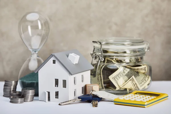 Hausmodell auf weißem Tisch mit Sanduhr, Taschenrechner, Geld und Schlüssel, Immobilienkonzept — Stockfoto