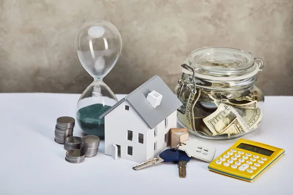 Modelo de casa con reloj de arena, calculadora, dinero y llaves, concepto de bienes raíces - foto de stock