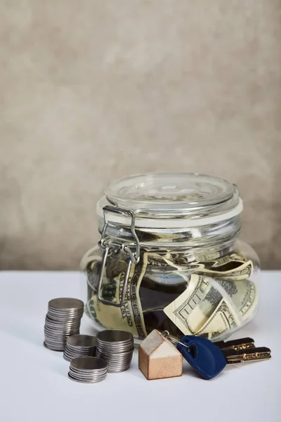 Spardose mit Dollarnoten, Schlüsseln und Silbermünzen auf weißem Tisch auf grauem Hintergrund, Immobilienkonzept — Stockfoto