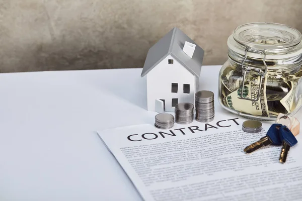 Hausmodell auf weißem Tisch mit Silbermünzen, Schlüsseln, Vertrag und Spardose, Immobilienkonzept — Stockfoto