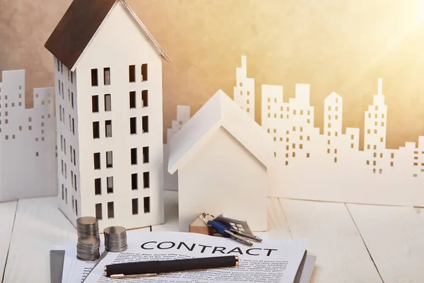Моделі будинків на білому дерев'яному столі з контрактом, монети і ключі біля білого паперу вирізане місто, концепція нерухомості — стокове фото