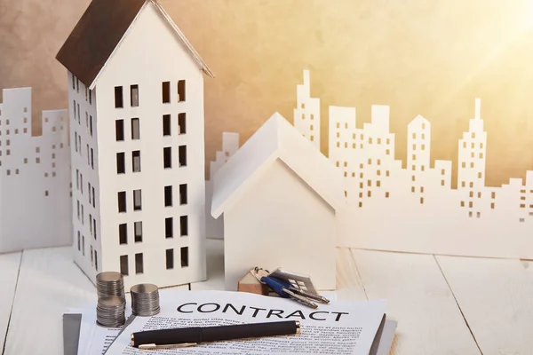 Houses models on white wooden table with contract, coins and keys near white paper cut city, real estate concept — Stock Photo