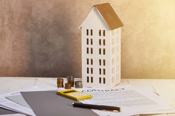 Hausmodell in der Nähe von Münzen, Taschenrechner, Stift und Vertrag auf dem Tisch mit Sonnenlicht, Immobilienkonzept — Stockfoto