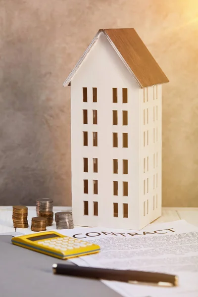 Maison modèle près de pièces, calculatrice et contrat sur table avec lumière du soleil, concept immobilier — Photo de stock