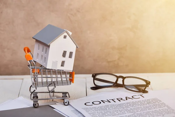 House model in small shopping cart on white wooden table with contract and glasses in sunlight, real estate concept — Stock Photo