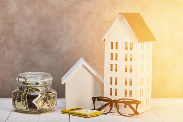 Casas modelos en mesa de madera blanca con gafas, calculadora y caja de dinero en sol, concepto de bienes raíces - foto de stock