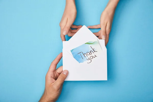 Cropped view of man giving greeting card with thank you lettering in envelope to woman on blue background — Stock Photo