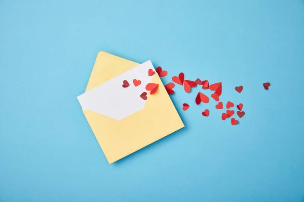 Желтый конверт с пустой белой картой и бумагой вырезать сердца на синем фоне — стоковое фото