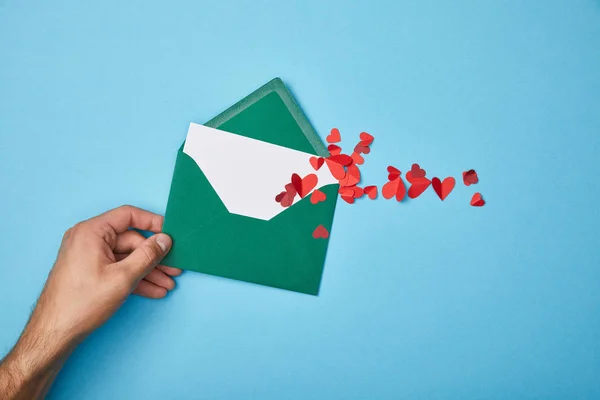 Vue recadrée de l'homme tenant une enveloppe verte avec une carte blanche vierge et des coeurs rouges coupés en papier sur fond bleu — Photo de stock