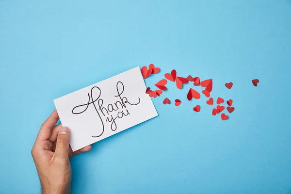 Cropped view of man holding white card with thank you lettering and red paper hearts on blue background — Stock Photo
