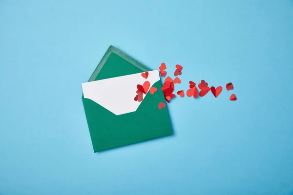 Sobre verde con tarjeta blanca en blanco y corazones de papel rojo sobre fondo azul - foto de stock