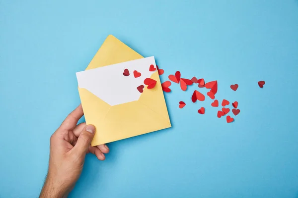 Visão recortada do homem segurando envelope amarelo com cartão branco em branco e corações de papel vermelho no fundo azul — Fotografia de Stock