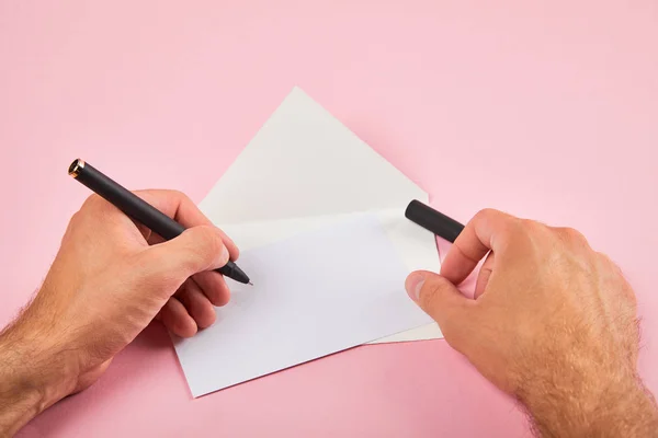 Ausgeschnittene Ansicht eines Mannes, der mit Stift auf weißer leerer Karte in der Nähe eines Umschlags auf rosa Hintergrund schreibt — Stockfoto