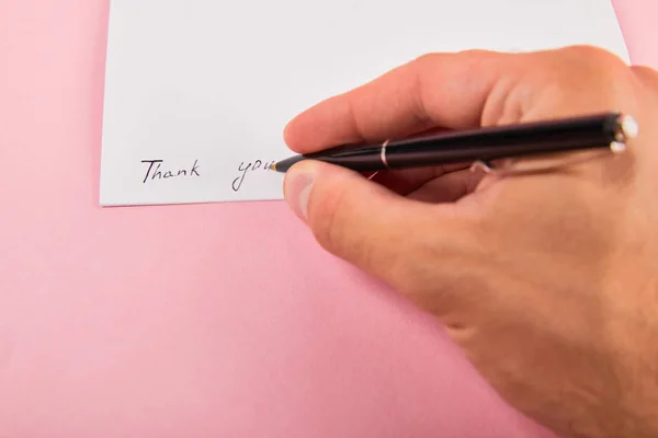 Recortado vista del hombre escribiendo con pluma gracias letras en la tarjeta blanca sobre fondo rosa - foto de stock