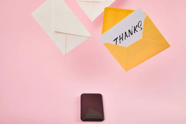 Yellow envelope with white card with thanks lettering near letters and smartphone with blank screen on pink background — Stock Photo