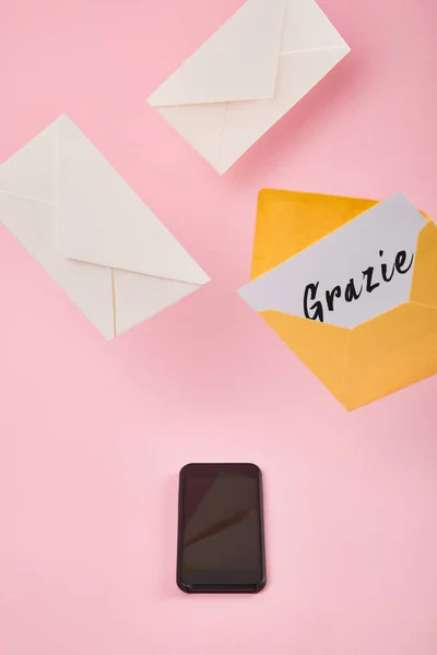 Yellow envelope with white card with grazie lettering near letters and smartphone with blank screen on pink background — Stock Photo