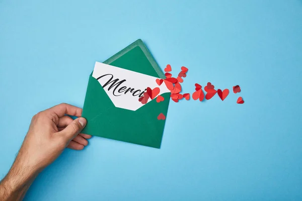 Cropped view of man holding green envelope with merci lettering on white card and paper cut red hearts on blue background — Stock Photo