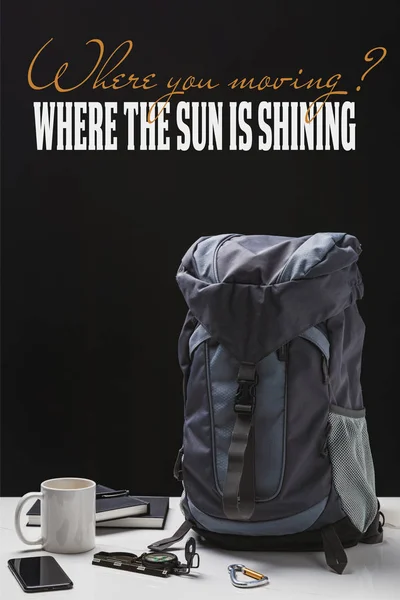 Backpack, cup, notebooks, smartphone and trekking equipment isolated on black with where you moving question and where the sun is shining answer — Stock Photo