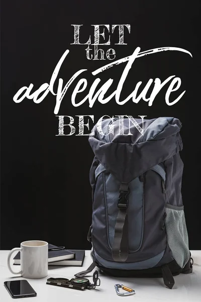 Mochila, taza, cuadernos, teléfono inteligente y equipo de trekking aislado en negro con dejar que la aventura comience a escribir - foto de stock