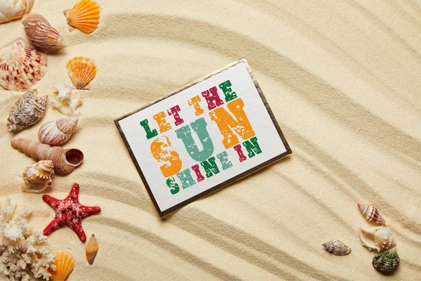 Top view of card with let the sun shine in lettering near seashells, red starfish and corals on sandy beach — Stock Photo