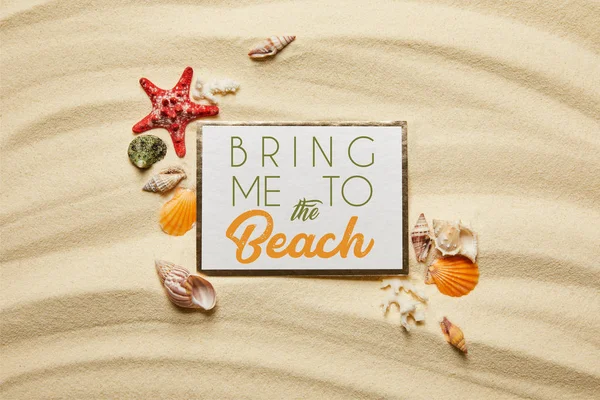 Top view of placard with bring me to the beach lettering near seashells, starfish and white corals on sandy beach — Stock Photo