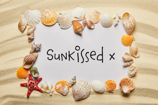 Marco de conchas marinas cerca de pancarta con letras besadas por el sol en la playa de arena - foto de stock