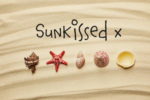 Puesta plana de conchas marinas y estrellas de mar rojas en la playa de arena en verano con letras besadas por el sol - foto de stock