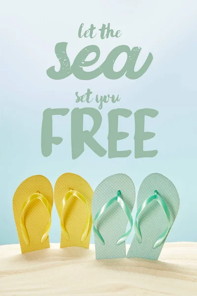 Summer yellow and blue flip flops in golden sand isolated on blue with let the sea set you free lettering — Stock Photo