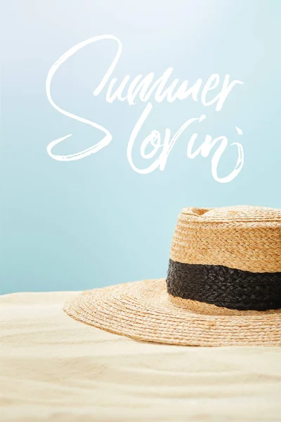 Enfoque selectivo de sombrero de paja sobre arena dorada en verano aislado en azul con letras de verano lovin - foto de stock