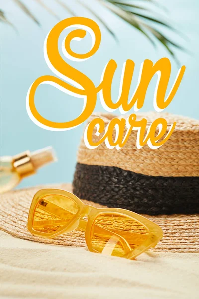 Gafas de sol cerca de sombrero de paja y botella con aceite bronceador en la playa de arena aislada en azul con ilustración de cuidado del sol - foto de stock