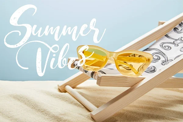 Yellow stylish sunglasses on deck chair on sand and blue background with summer vibes lettering — Stock Photo
