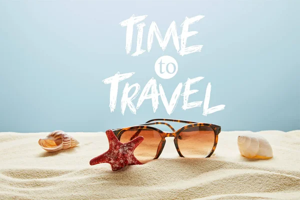 Óculos de sol castanhos elegantes na areia com conchas e estrelas do mar em fundo azul com tempo para viajar lettering — Fotografia de Stock