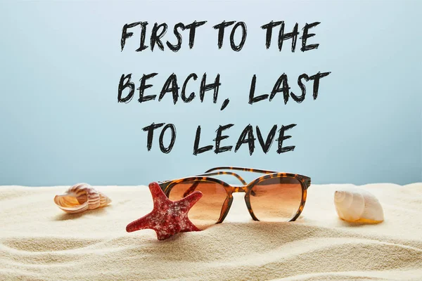 Gafas de sol de estilo marrón sobre arena con conchas marinas y estrellas de mar sobre fondo azul con primera a la playa, últimas en dejar letras - foto de stock