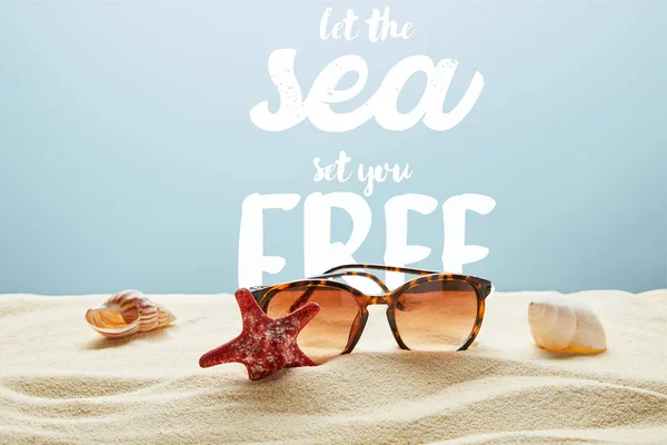 Brown stylish sunglasses on sand with seashells and starfish on blue background with let the sea set you free lettering — Stock Photo