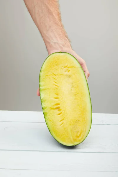 Cropped view of man holding ripe yellow watermelon half on wooden table isolated on grey — Stock Photo