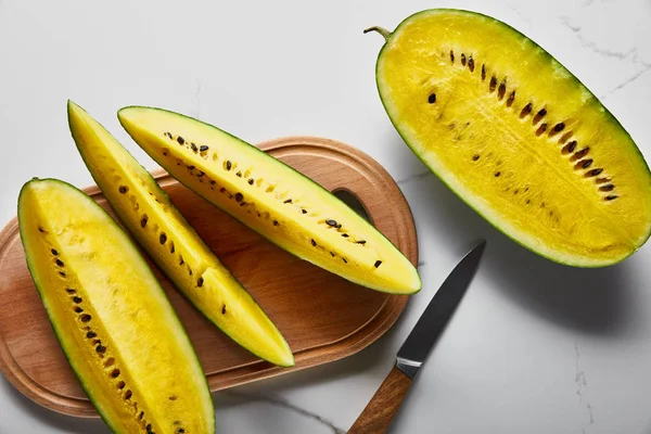 Vue du dessus de la délicieuse pastèque jaune exotique coupée avec des graines sur planche à découper en bois avec couteau sur la surface du marbre — Photo de stock